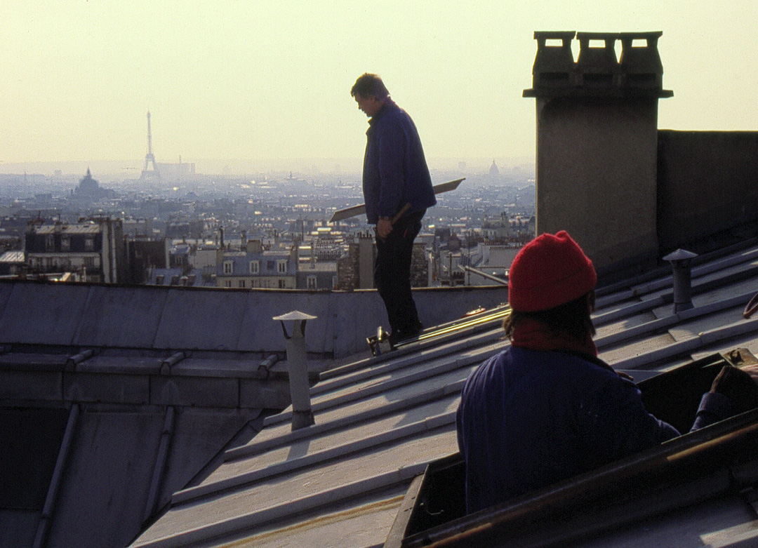 Two roofers work high above the streets of Paris, surrounded by zinc rooftops and chimneys. In the distance, the Eiffel Tower rises through the hazy skyline. The scene captures the balance, concentration, and sense of freedom that define life on the rooftops—like sailors working above an endless ocean of buildings.